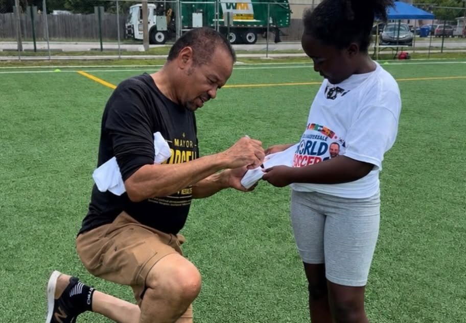 Mayor Borgelin kneeling on a soccer field, signing a jersey for a young girl at a youth soccer event